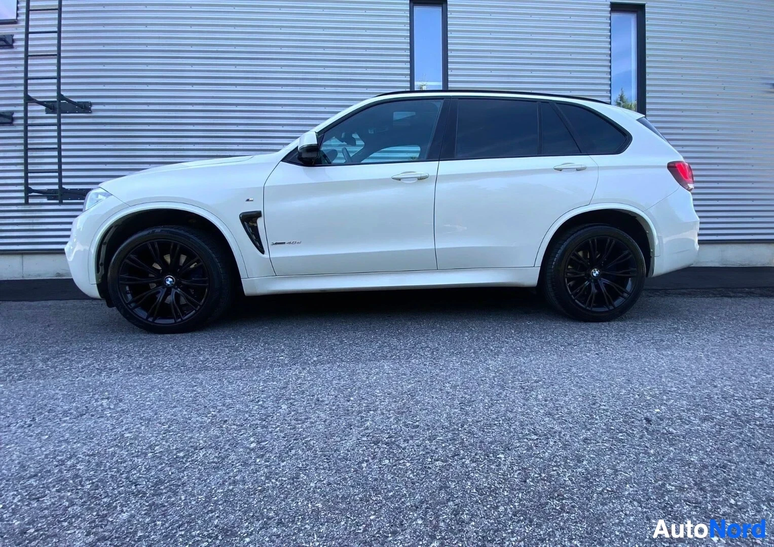 Sivulta: Valkoinen BMW X5 F15 -mallin katumaisen talon seinää vasten, näkyy vasemmasta puolesta, mustat rengaskierrokset.