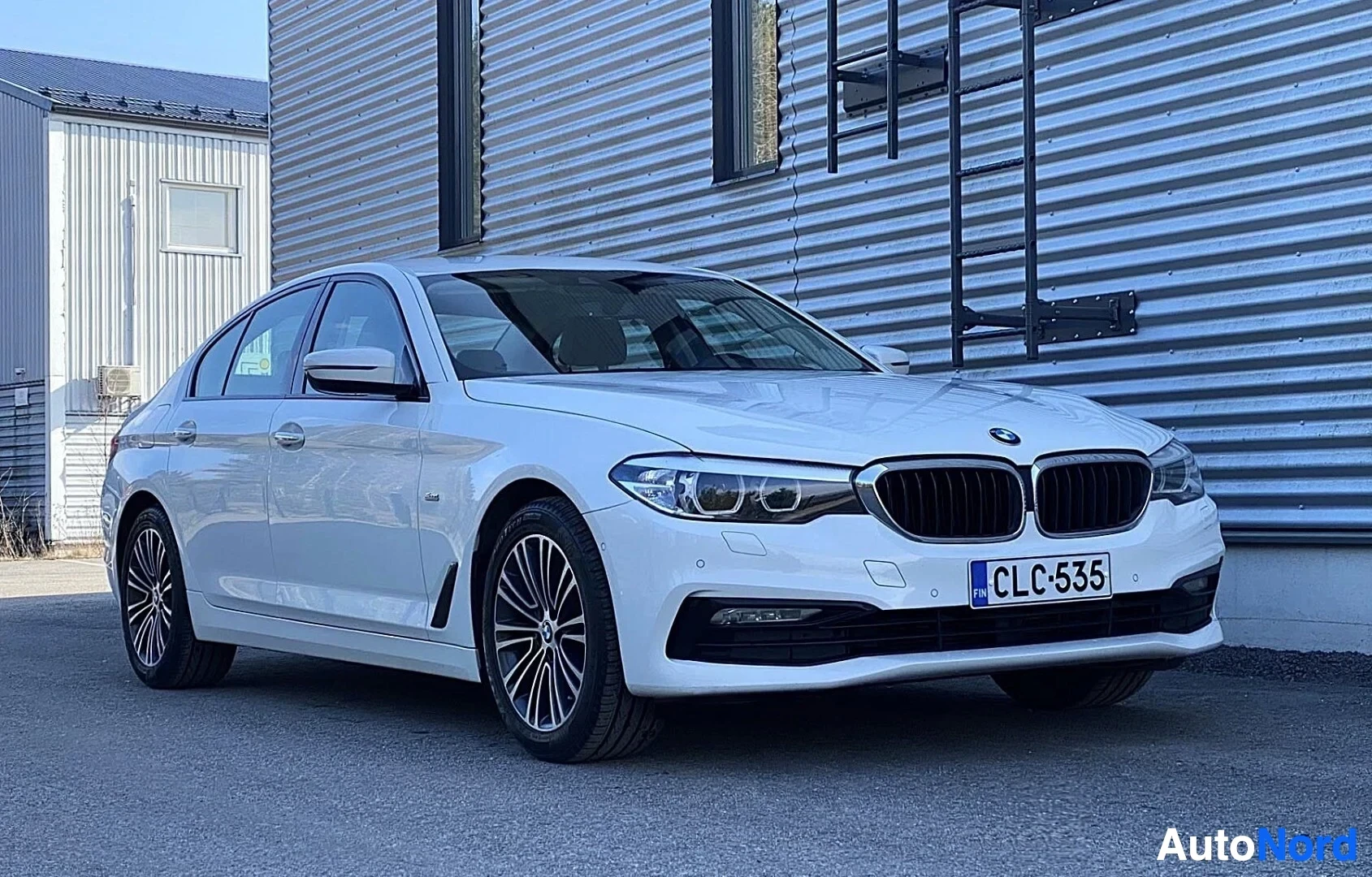 Etuviisto, nokka vasemmalle: Valkoinen BMW-sedan näkymässä kolmen neljänneksen etulta vasemmalle, teräsvärisellä rakennuksella taustalla. Rekisterikyltti näkyvissä.