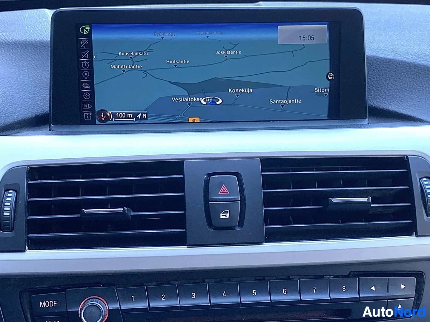 Yksityiskohta: BMW navigaattonäytön lähikuva. Näyttö näyttää karttaa Keski-Suomesta ajanotilla Vessilahti, Konekuja ja Sitom. Aika 15:05. Alla ilmastointi-ohjaimet ja MODE-nap