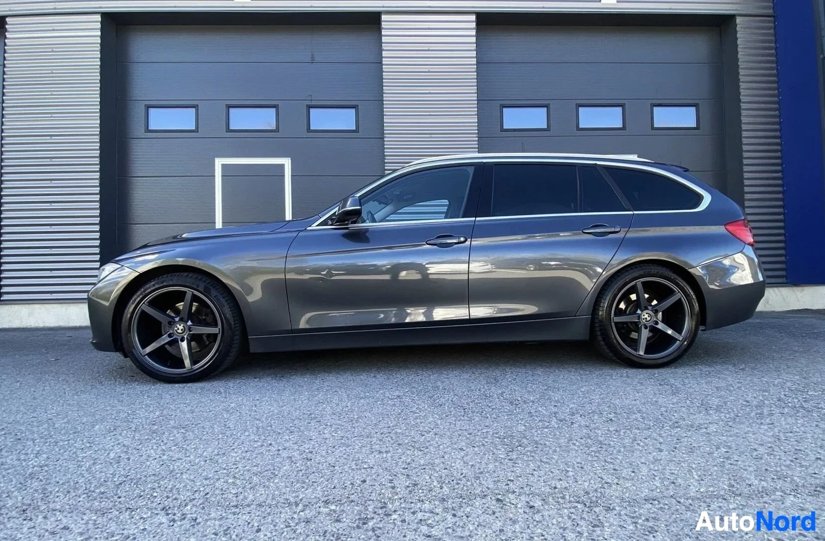 Sivulta: Harmaa BMW-farmari sivusta oikealle, tummansinisessä hallissa.