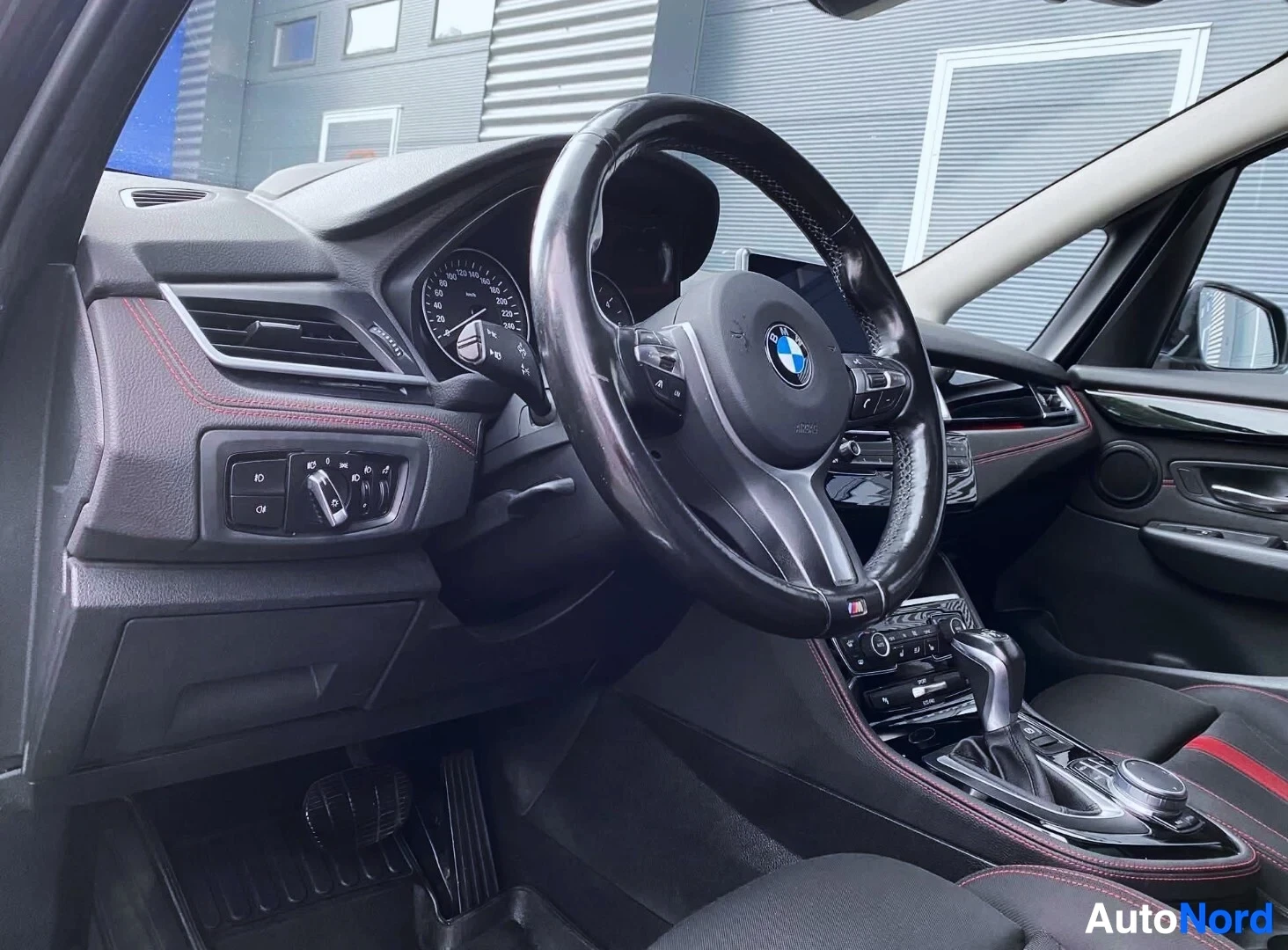 Ohjaamo: Kuljettajan puoleinen näkymä sisustaan, BMW-logo ohjauspyörässä, mustat istuin punaisilla yksityiskohdilla, valkoiset vanteet näkyvissä