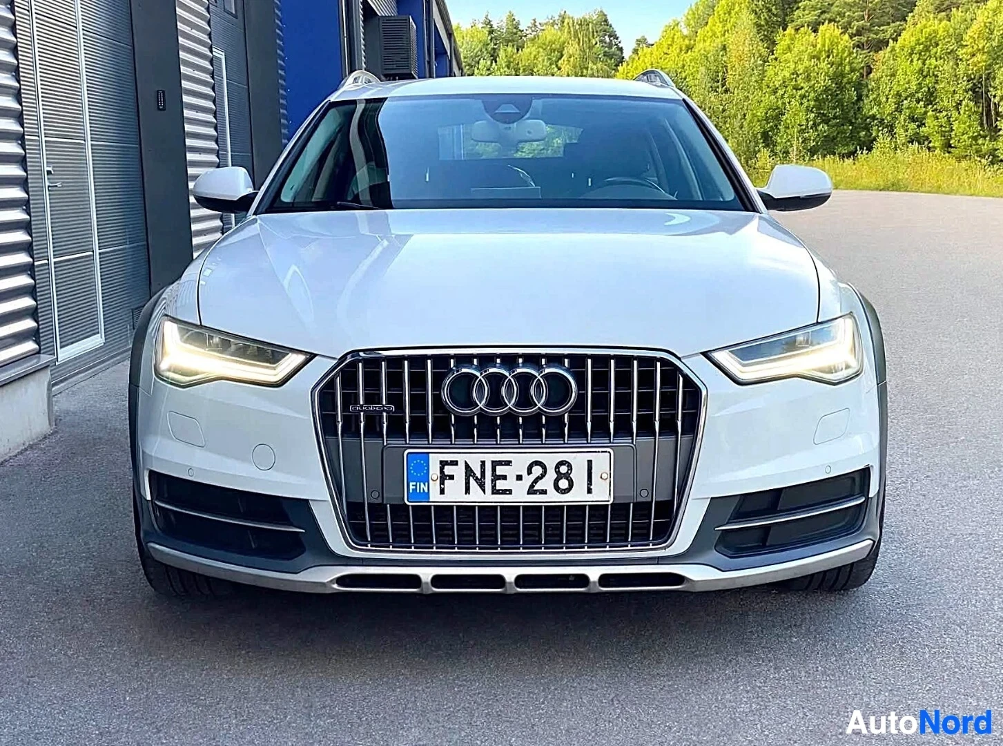 Edestä: Valkoinen Audi-farmari kuvattuna suoraan edestä, pysäköitynä teollisuusrakennusten eteen.