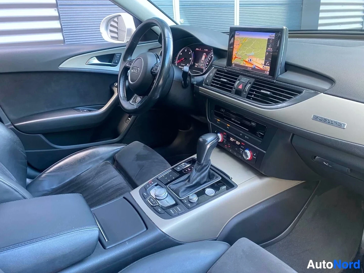 Ohjaamo: Auton sisäkuva: musta monikäyttöinen ohjauspyörä Audi-logolla, digitaalinen mittaristo, infotainment-näyttö kartalla ja liikennetiedoilla, ilmastointi-ohjaimet,