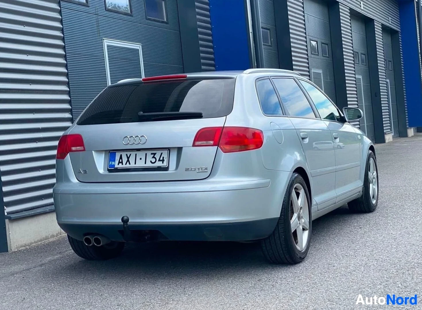 Takaviisto: Hopeinen viistoperä takakulmasta oikealle, pysäköity teollisuusrakennusten eteen. Näkyy punainen takavalo, Audi-logo ja rekisterikylpi