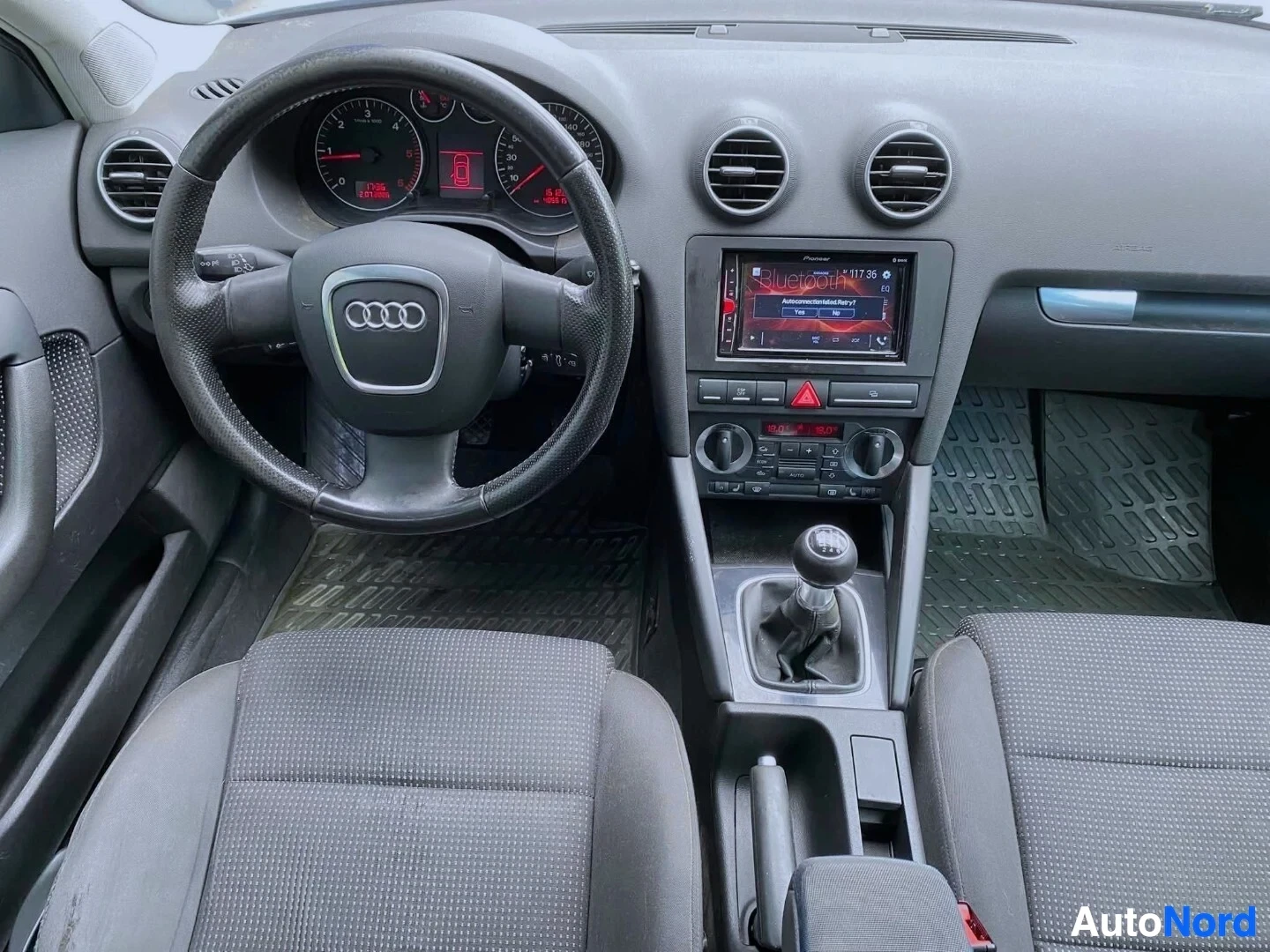 Etuistuimet: Kojelauta suoraan, Audi-logo ohjauspyörässä, instrumentit ja näyttö näkyvillä, harmaa verhoilu etupenkillä, automaattivaihteisto keskikonsolissa.