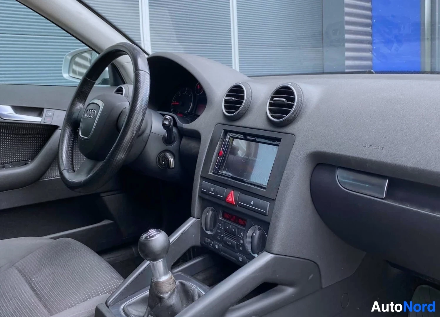 Etuistuimet: Kojelauta sivusta kuvattuna, Audi-logo ohjauspyörässä, näyttö keskellä, harmaa panelointi, kolmivaihteinen vaihdevipu.