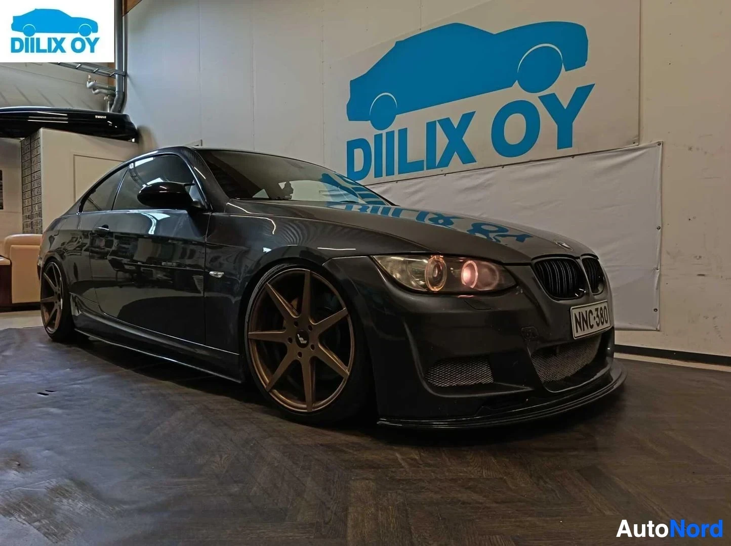 Etuviisto, nokka vasemmalle: Musta BMW 335 E92 coupe vasemmalla viistolla, kulta-/pronssinväriset vanteet, punaiset takavalosuulakkeet, näkyvissä rekisterikyltti