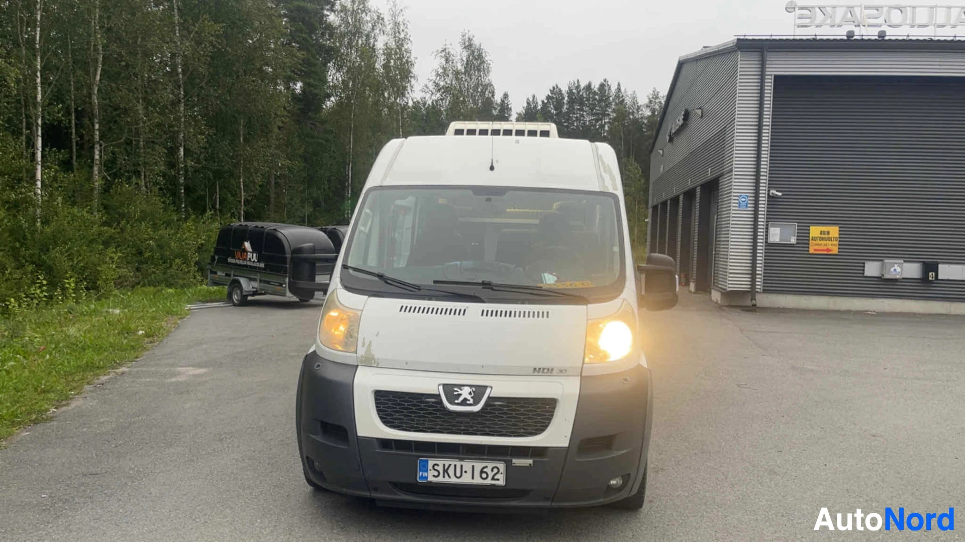 Edestä: Valkoinen Peugeot-pakettiauton etunäkymä edestä, rekisterinumero näkyvissä, puut taustalla