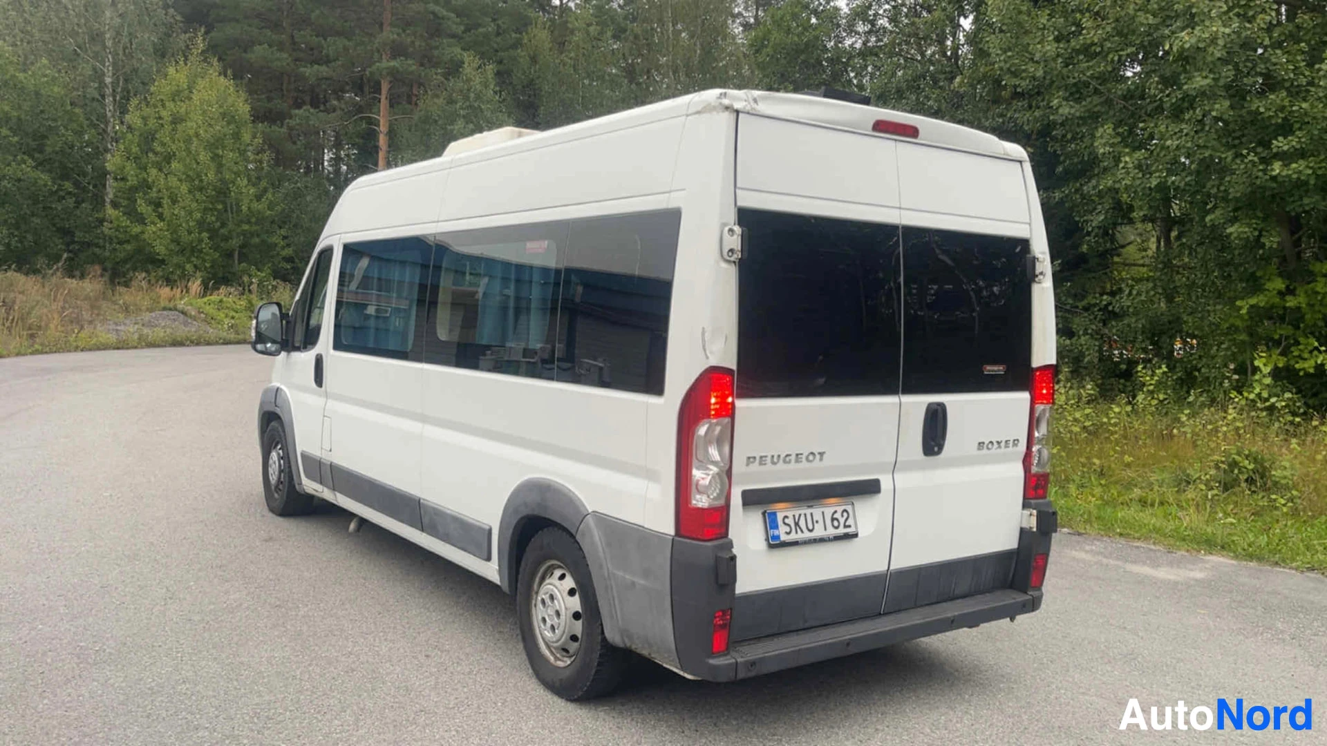 Takaviisto: Valkoinen Peugeot Boxer -pakettiauto näkymässä takaa. Rekisterikilvessä. Punaisia takavalojas ja mustat raidat sivuilla. Pysäköity sorateille metsän vie