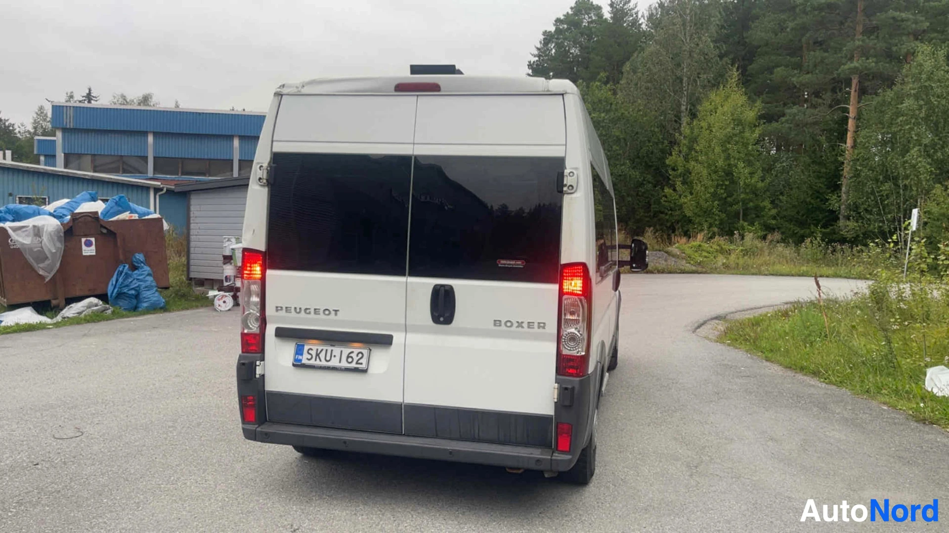 Takaa: Valkoinen Peugeot Boxer -pakettiauto näkymässä takaa. Rekisterikilvessä. Takavalojas punaisella valaistuksella. Sininen keräysrakennus vasemmalla tausta