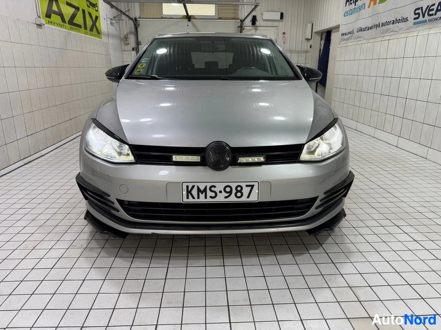 Edestä: Hopeinen viisioviinen Volkswagen Golf henkilöauto suoraan etuosasta kuvattuna hallin sisällä. Rekisterikilvessä. LED-sisääntulopalkit otsikossa. Musta r