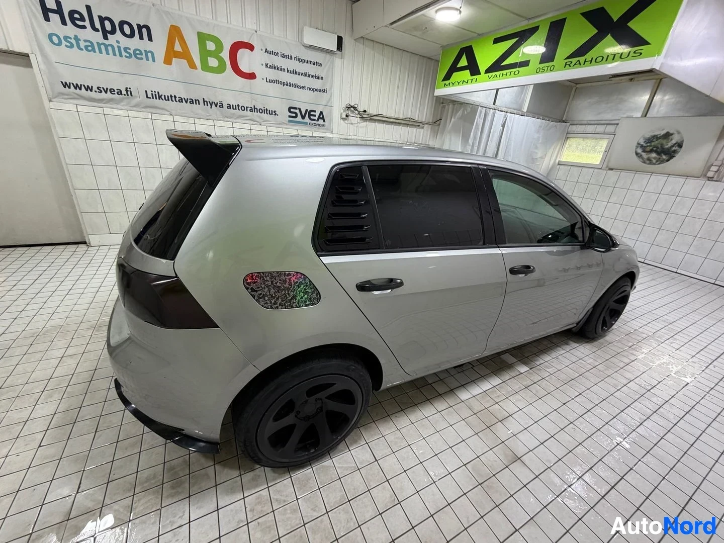 Takaviisto: Valkoinen Volkswagen Golf seisoo sisätiloissa, katsottuna sivulta ja takaosasta. Rekisterikilpi näkyy osittain.