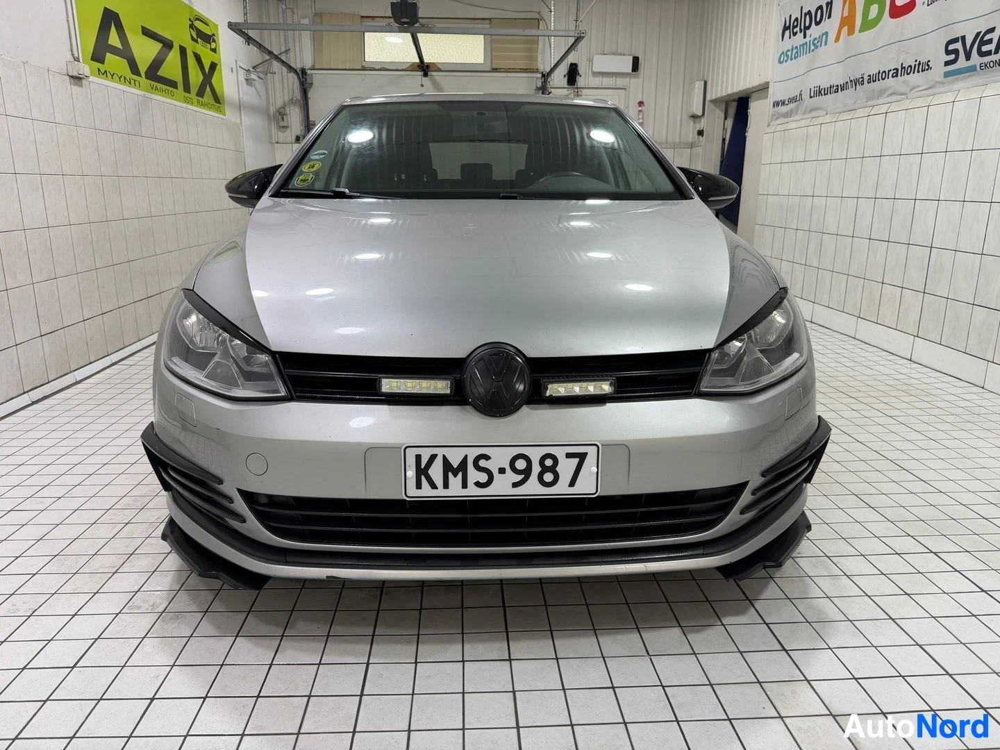 Edestä: Valkoinen Volkswagen Golf suoraan edestä kuvattuna sisätiloissa. Rekisterikilpi ja VW-logo grillissä näkyvät