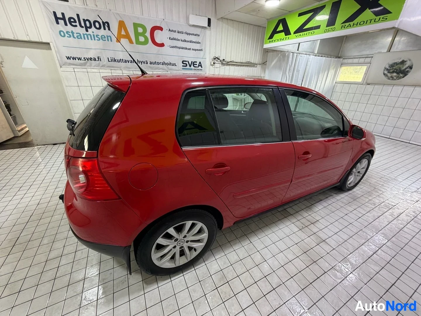 Sivulta: Punainen Volkswagen Golf seisoo sisätiloissa sivusta kuvattuna oikealle puolelle. Avoimet ovet, viisinkertainen paikka ja valurenkaat ovat näkyvissä. Rekisterik