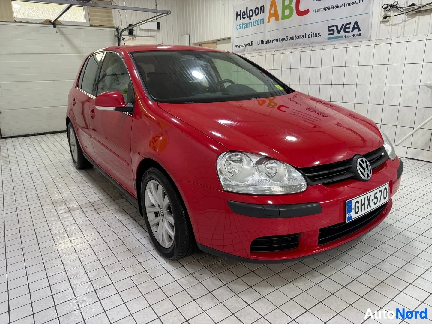Etuviisto, nokka vasemmalle: Punainen Volkswagen Golf seisoo sisätiloissa edestä vasemmalle kulmasta kuvattuna. Etupää, vasen sivu ja pyörät näkyvät. Valurenkaat ja kaksoisajovalot