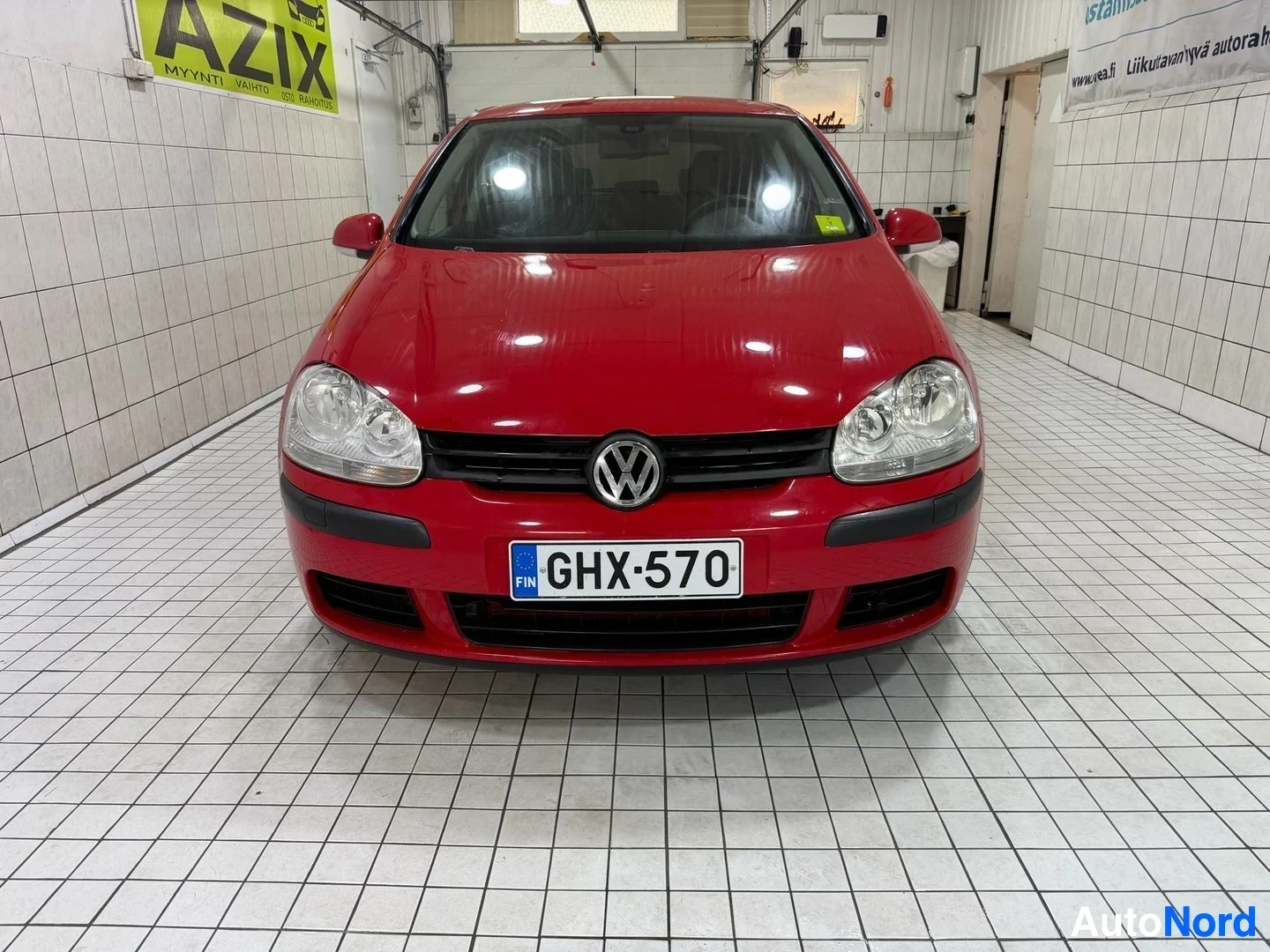 Edestä: Punainen Volkswagen Golf seisoo sisätiloissa edestä kuvattuna. Etupuskuri, kaksoisajovalot, Volkswagen -merkki ja rekisterikilpi ovat näkyvissä. Auto on