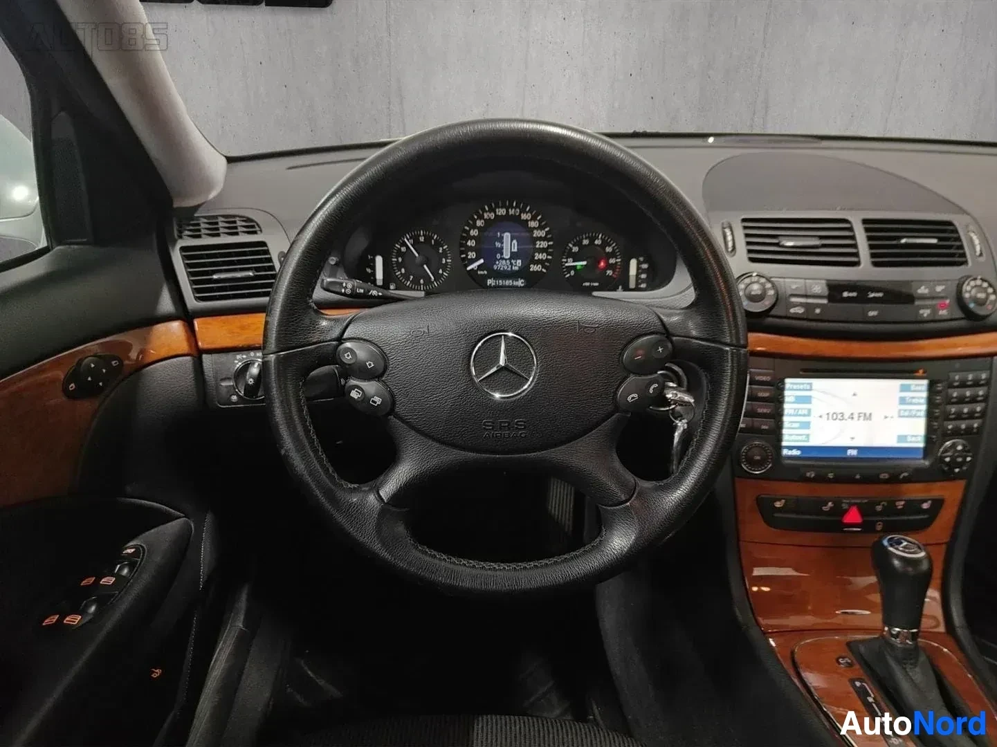 Yksityiskohta: Lähikuva Mercedes-Benz rattista ja mittarista: musta nahkaryhtävä ratti kahvalla, kotiintulo-merkinnöillä ja oranssi puurengas, yläpuolella ajomittari joka näyt