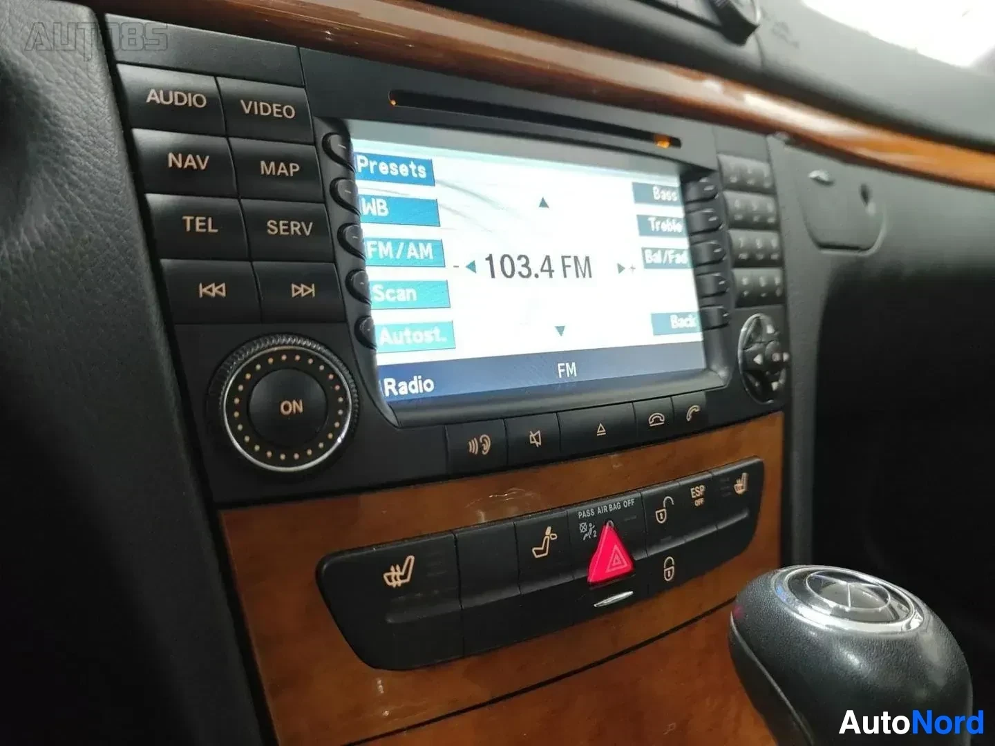 Yksityiskohta: Navigaatiolaitteen näyttö radiokanavalla, navigointivalikko näkyvillä sekä ohjaimen painikkeet ja puupanelointi.