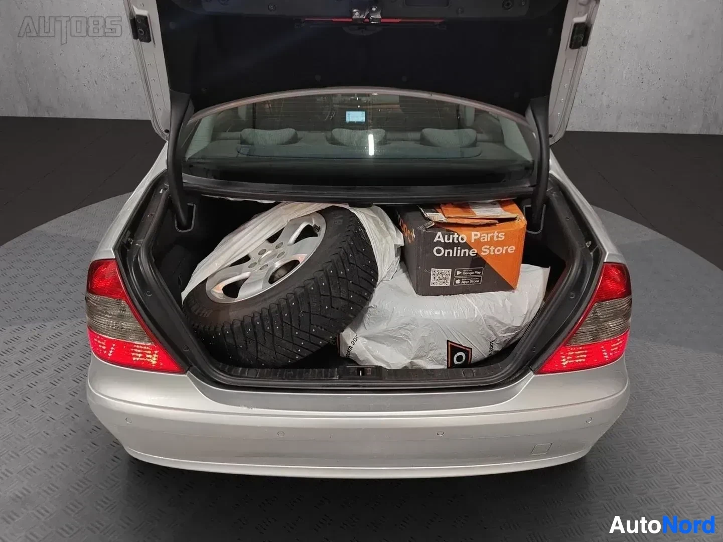 Tavaratila: Auton avoin tavaratila, jossa on talvisarja (rengas ja kiekko) sekä oranssin väriset Auto Parts Online Store -pakkauksissa olevia osia.