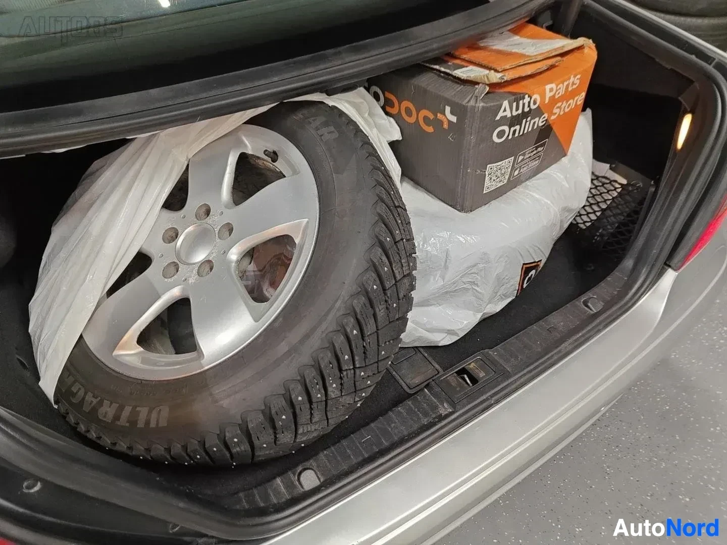 Tavaratila: Auton takakontin sisällä varastoitu alumiinivanne ja talvirengas sekä Auto Parts Online Store -merkinnällä varustettu paketti.