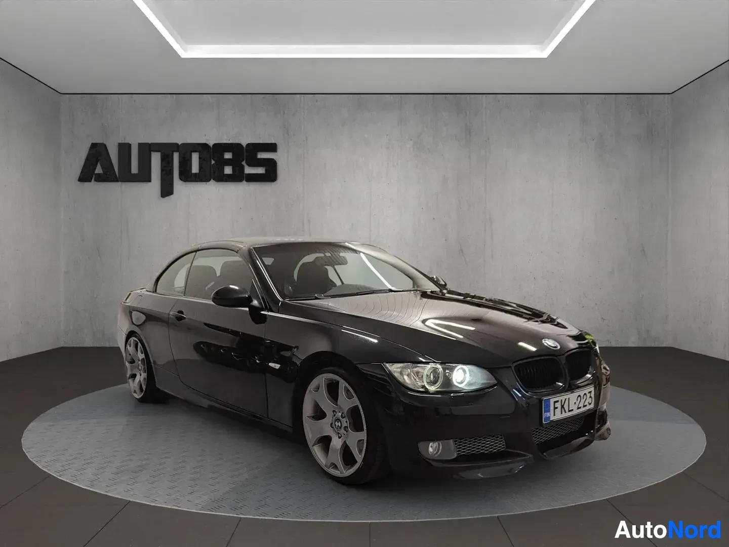 Sivulta: Musta BMW coupe näkyy oikealta sivulla suljetulla katolla, valkoiset alumiinivanteet, harmaalla lattialla.