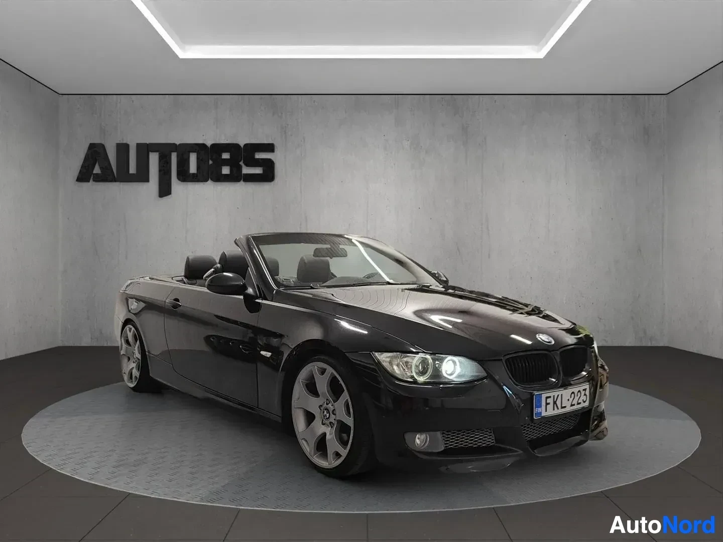 Sivulta: Musta BMW avoauto näkyy vasemmalta sivusta avoimella katolla, valkoiset alumiinivanteet, sisäpihan näyttämö.
