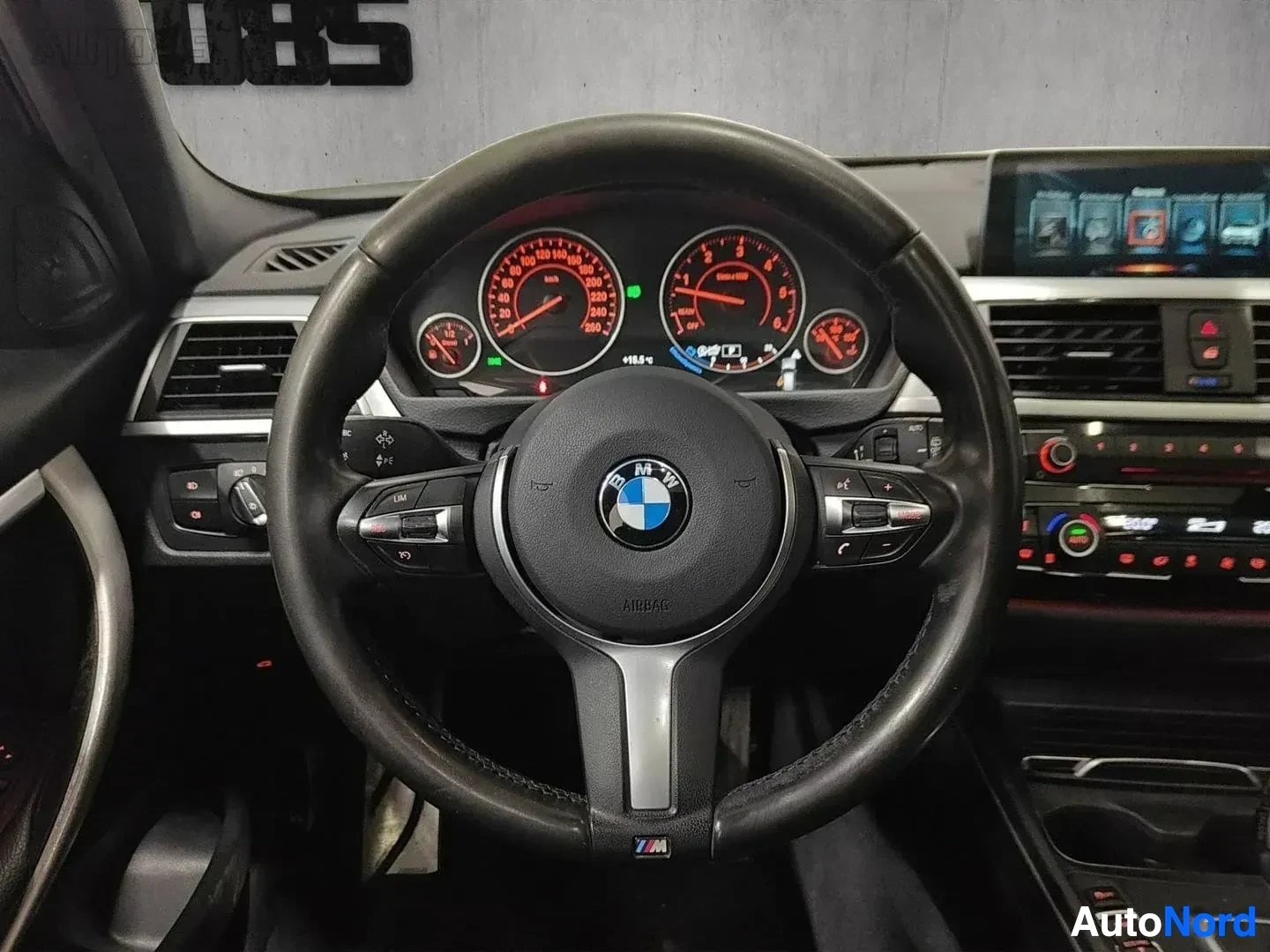 Yksityiskohta: Lähikuva BMW-auton ohjauspyörästä ja mittaristosta. Punaisten numeroiden mittaristo näyttää nopeuden ja moottorin kierroksia. Sininen BMW-merkki näkyy ohjauspyö