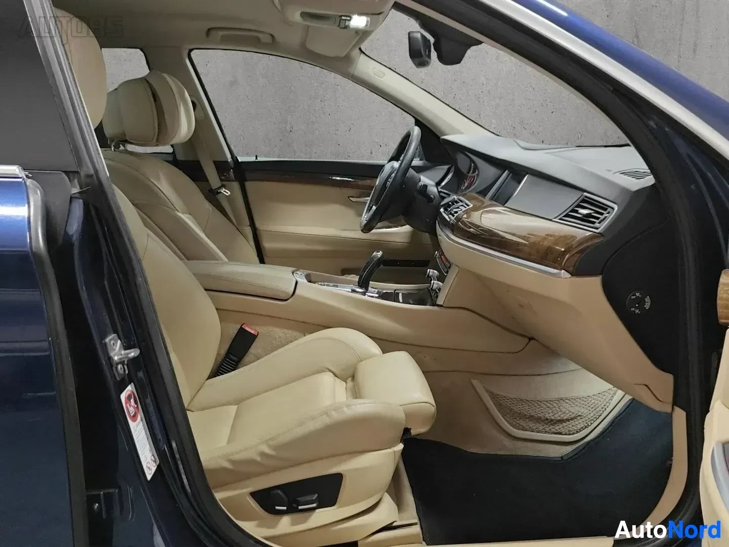 Etuistuimet: Auton etupaikat vasemmalta puolelta, beige nahkaverhoitus, ruskea puu-kalusteilla varustettu kojelauta, mustavalkoinen ohjauspyörä, sininen auto ulkona