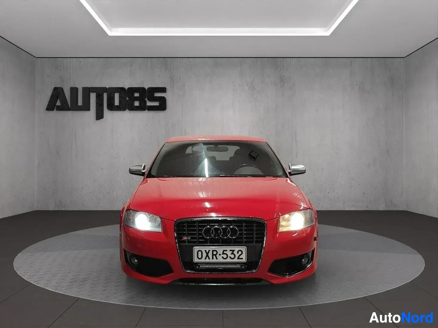 Edestä: Punainen Audi-auto suorasta edestä, xenon-valaisimet päällä, mustainen etumaski, valkoinen rekisterikyltti, showroom-valaistus.