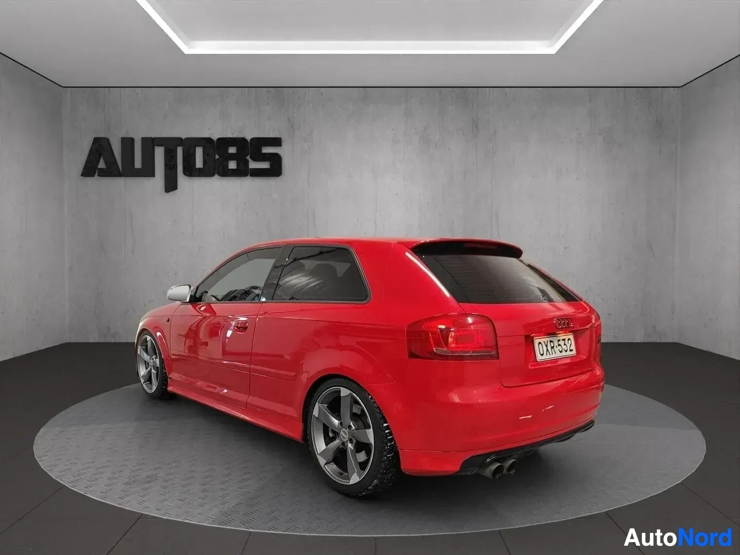 Takaviisto: Punainen Audi-auto näkyy takaa ja sivulta avoimella ovella studiossa.