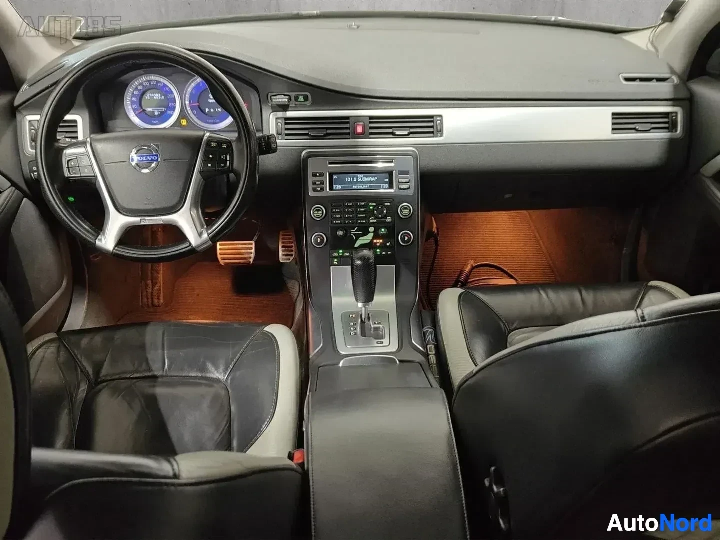 Ohjaamo: Volvo auton ohjaamotaulun näkymä edestä, nahkainen ratti, sininen valaistu mittaristo, puu-intarsia, automaattinen vaihteisto ja radio näkyvissä.