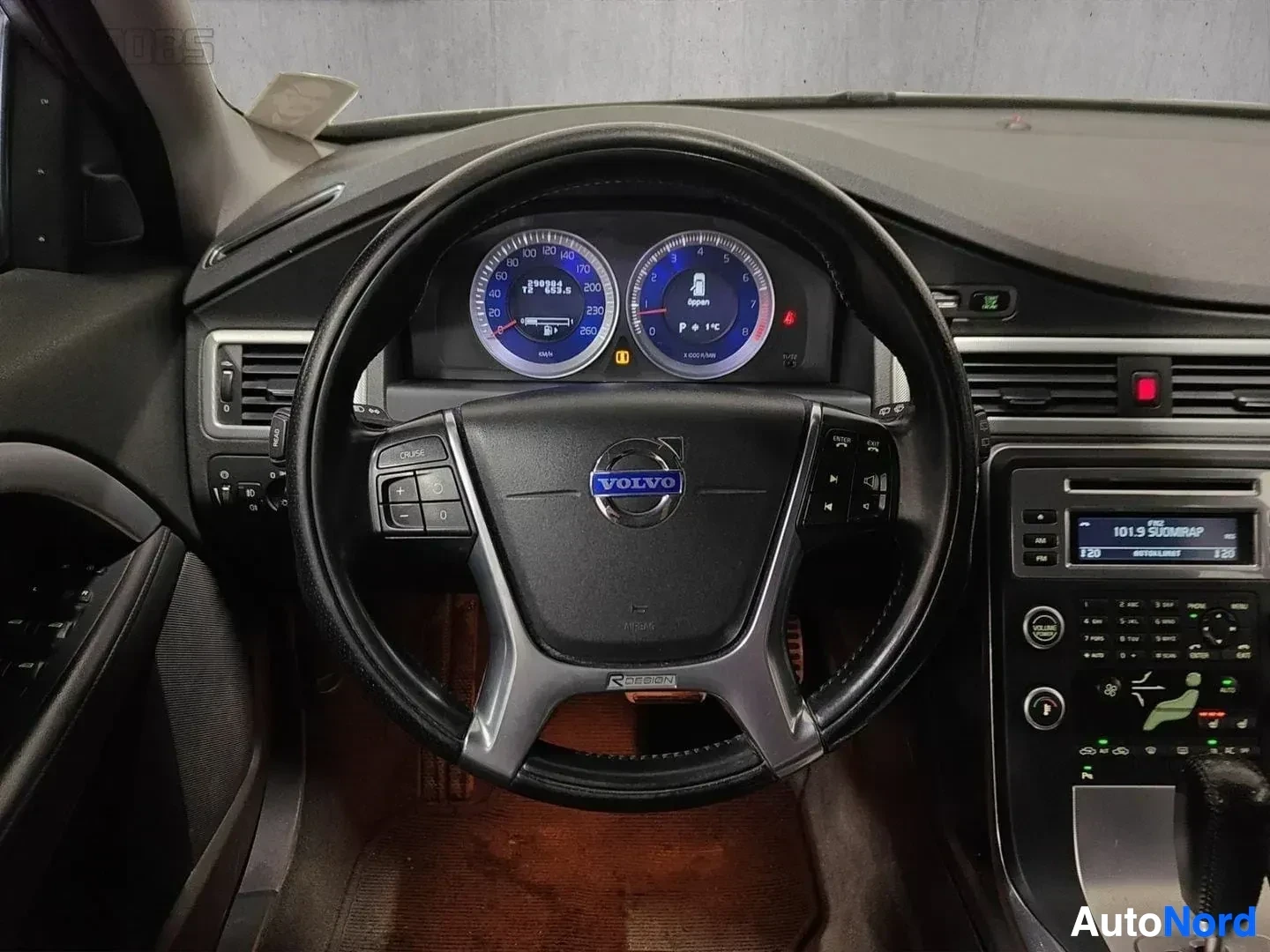 Ohjaamo: Volvo-merkkinen auto ohjaamosta katsottuna, nahkainen ratti 'R-DESIGN' merkinnällä, sininen valaistu mittaristo, puu-intarsia trimmi ja radio näkyvillä.