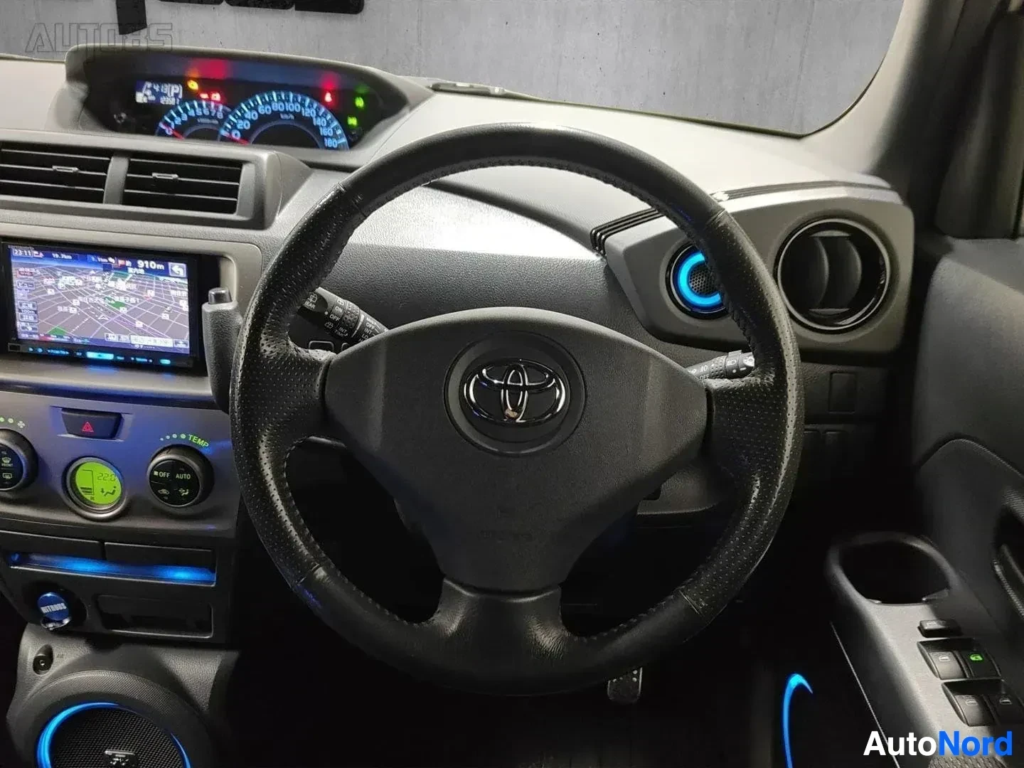 Ohjaamo: Kuljettajan näkökulma sisältäpäin: mustaksi verhoiltu ohjauspyörä Toyota-logolla, instrumenttipaneeli sinisin mittarein, navigaatiolaite vasemmalla ja ilmastoin
