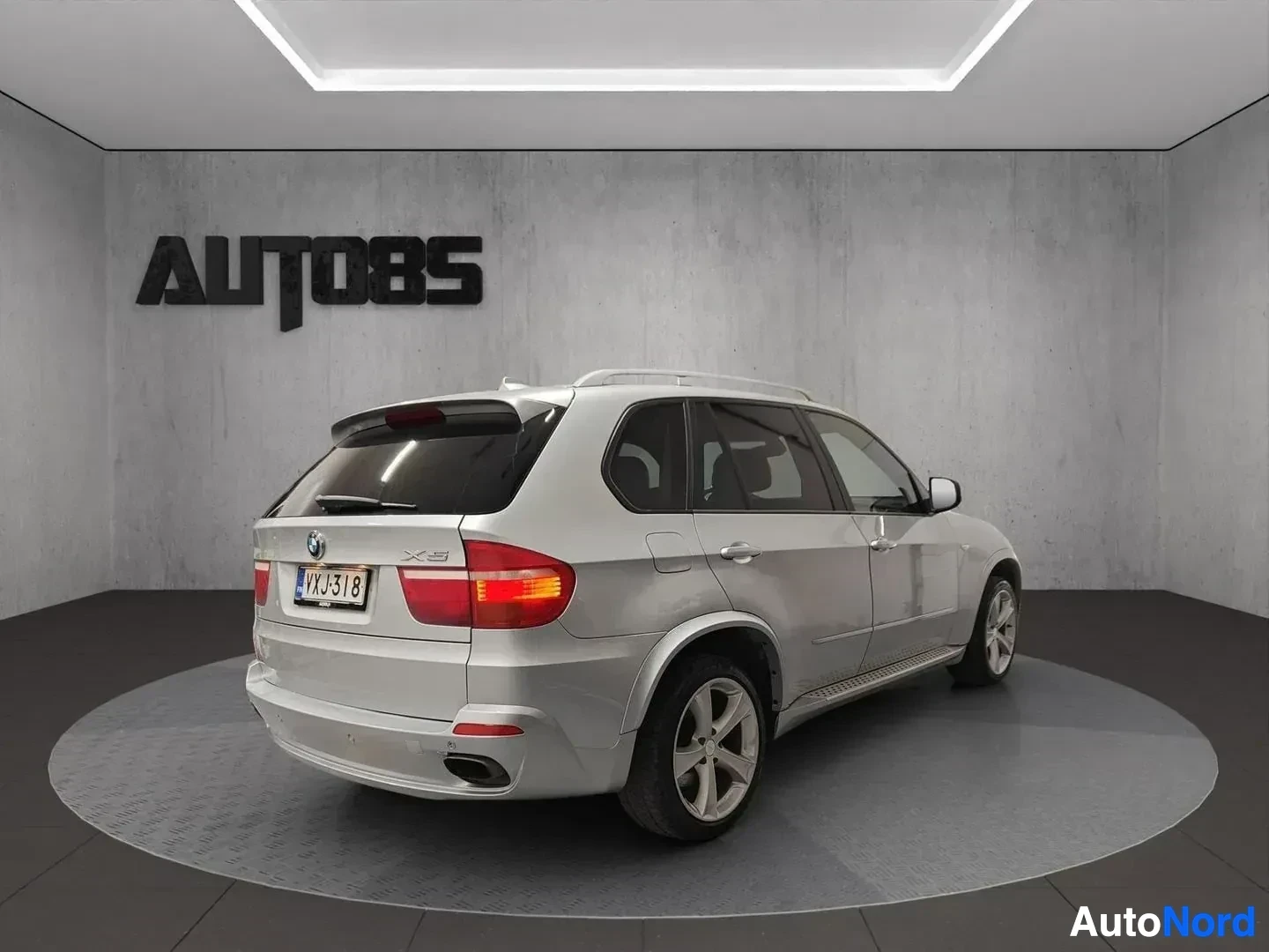 Takaviisto: Hopeanvärinen BMW X5 -maastoauto takaviistosta kuvattuna. Rekisterikilpi näkyy selvästi: vIV-J318. Auto seisoo studiossa.