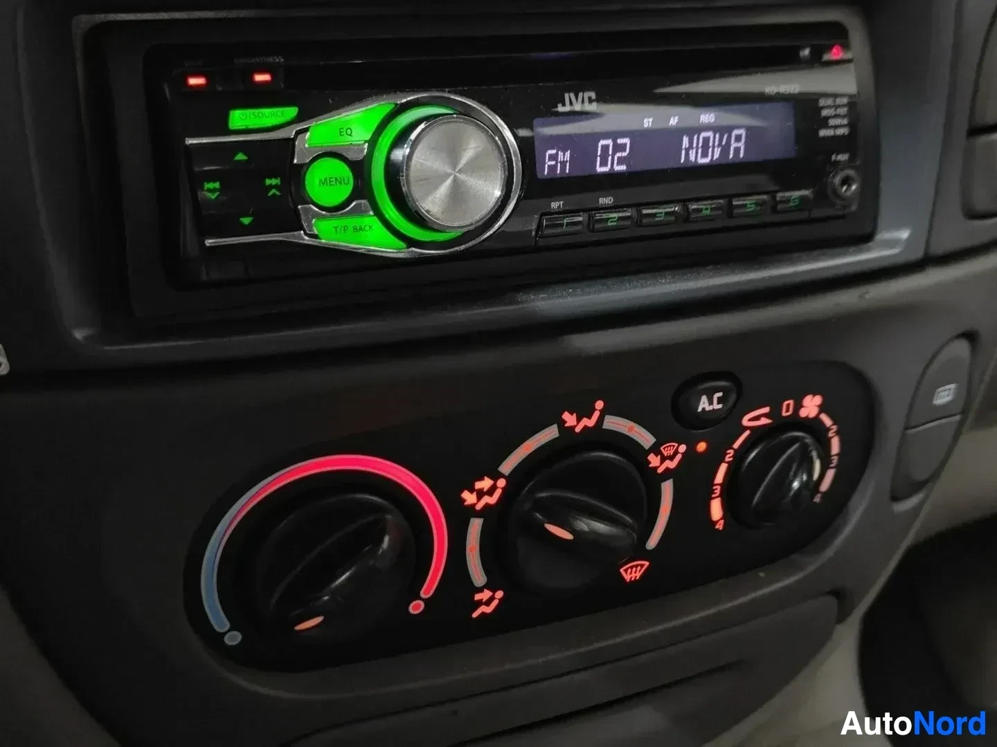 Yksityiskohta: Autoradio JVC-merkintä näkyvissä, vihreällä illuminaatiolla varustettu CD-soitinradio, ilmastoinnin ohjainpaneeli alla AC-napilla ja säätöpyörinä