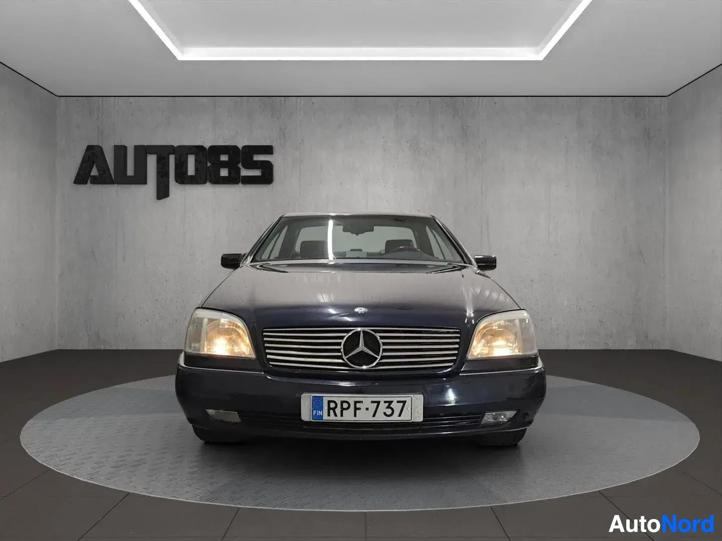 Edestä: Tummansininen Mercedes-Benz -luksusauto kuvattu suoraan edestä varustushallissa, rekisterikilpi näkyvissä.