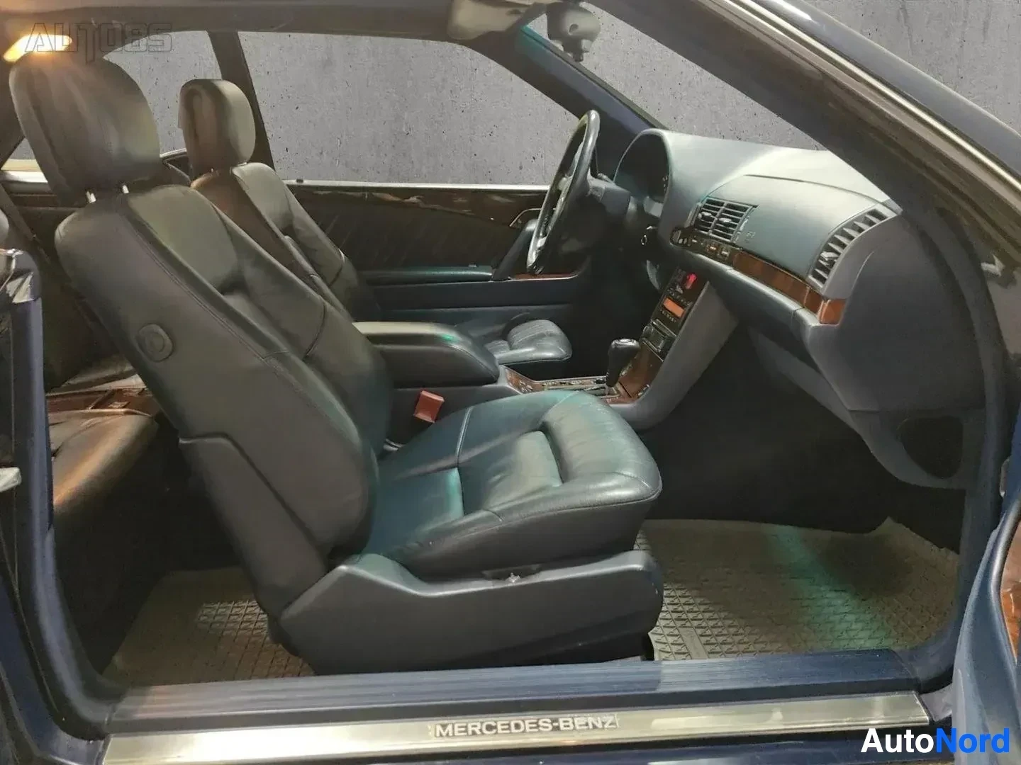 Etuistuimet: Avoimen oven kautta näkyvät etupaikat, nahkaverhoilu, puinen sisustuslevy, ohjauspyörä ja kojelauta, auton pohjan merkinnöissä lukee MERCEDES-BENZ.