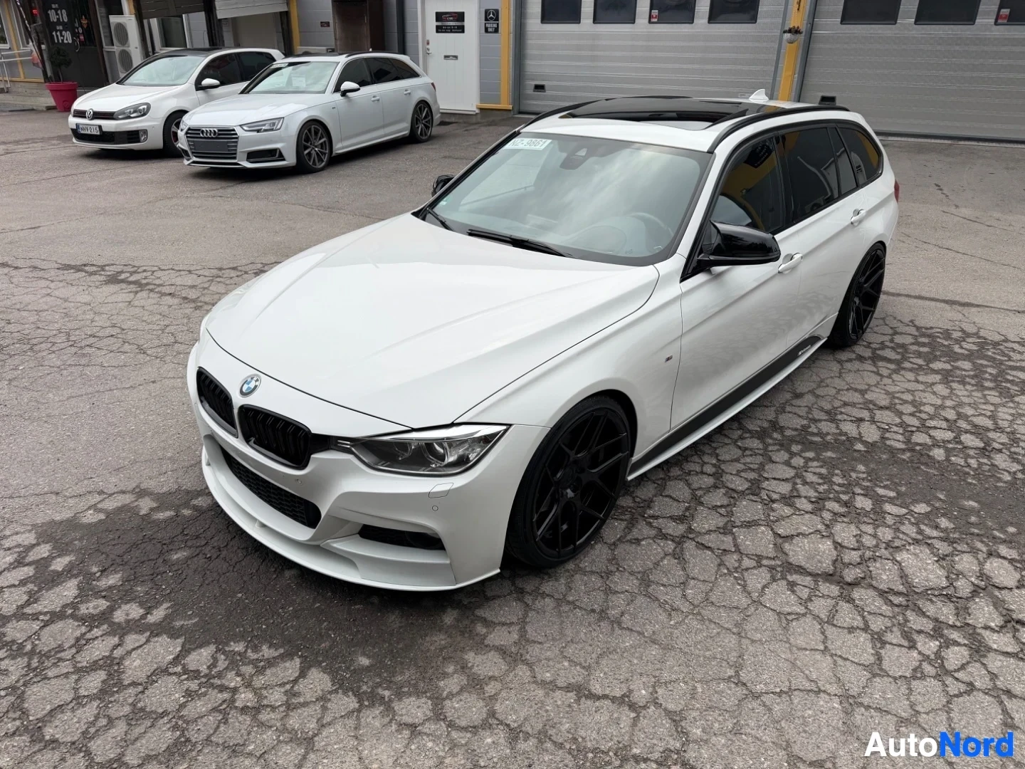 Valkoinen BMW farmari edestä oikealle kulmasta kuvattu, mustat vanteet, parkkipaikalla