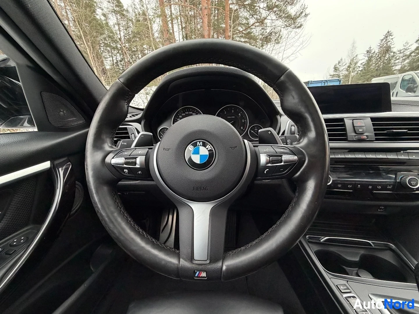 Yksityiskohta: Näkymä ohjausrenkaasta etupuolelta, BMW-logo ohjausrenkaan keskellä, M-striimi ohjausrenkaan alaosassa, mittarit ja keskikonsoli näkyvissä taustalla