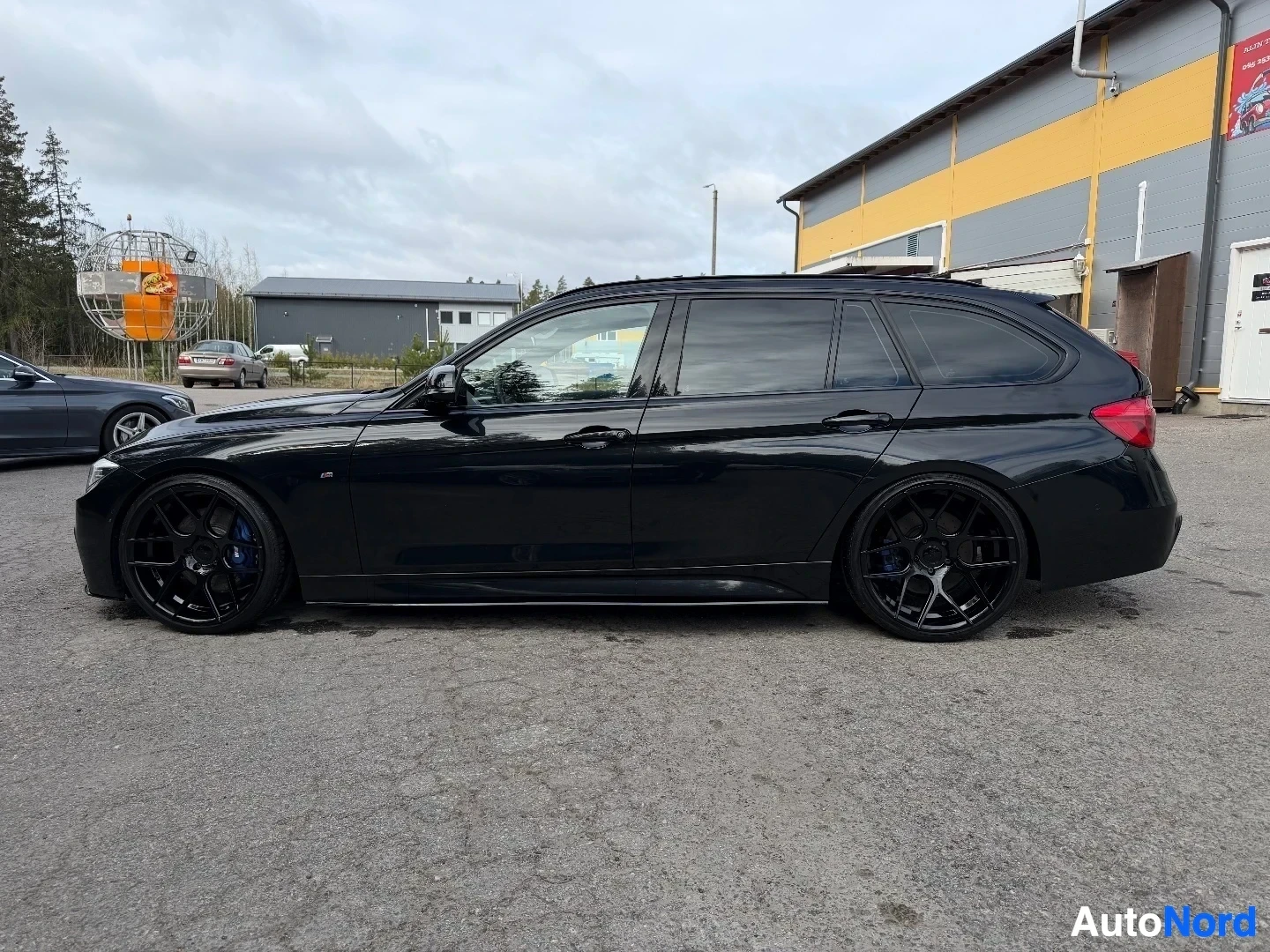 Musta BMW farmari näkyy sivusta ajoneuvon kanssa teollisuusrakennuksella.