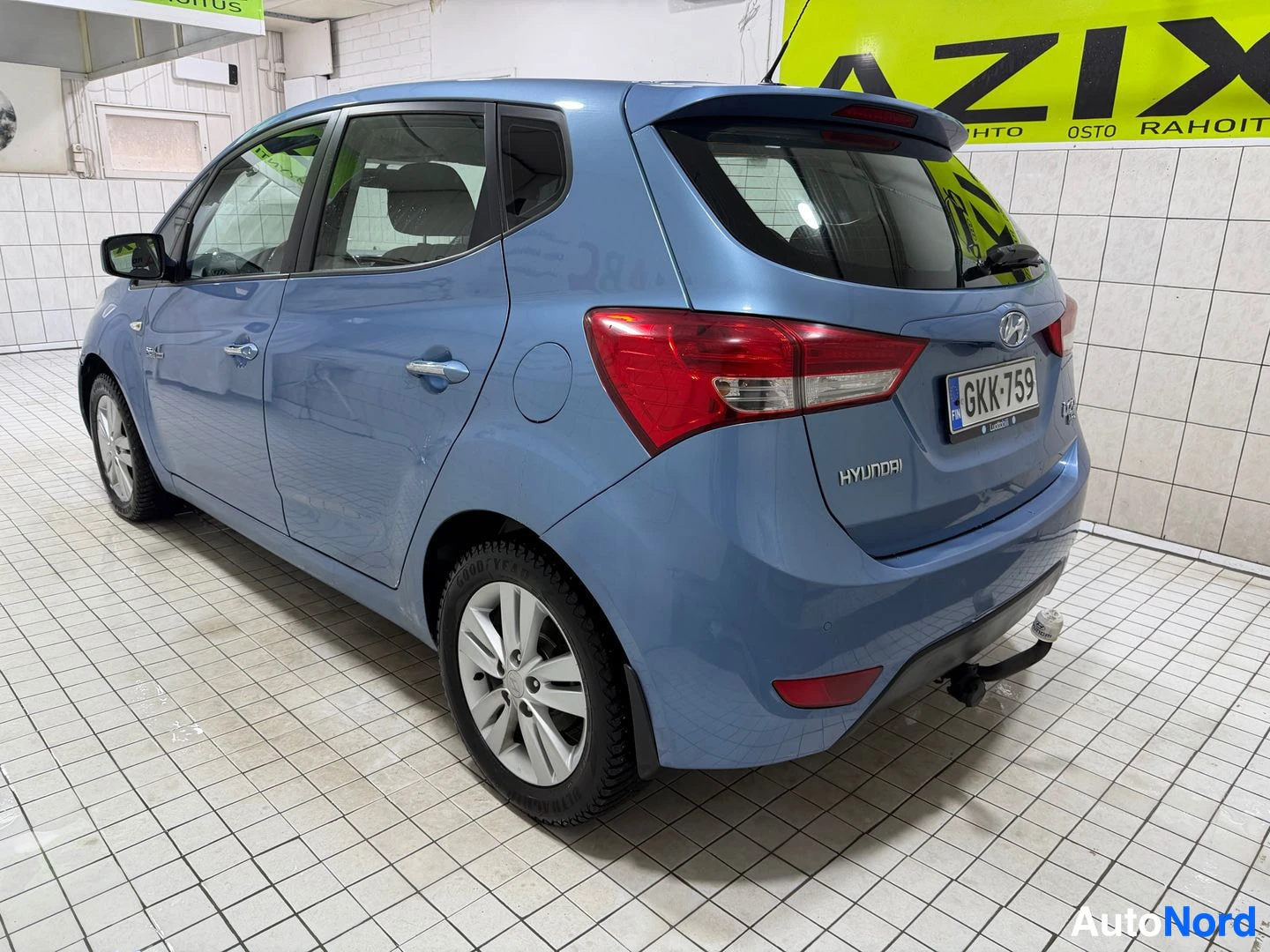 Takaviisto: Sininen Hyundai ix20 takaa ja oikealta viistosti kuvattuna. Autossa on peräkoukku ja takalasinpyyhin. Rekisterikilvessä. Ikkunoissa näkyy heijastuksia.
