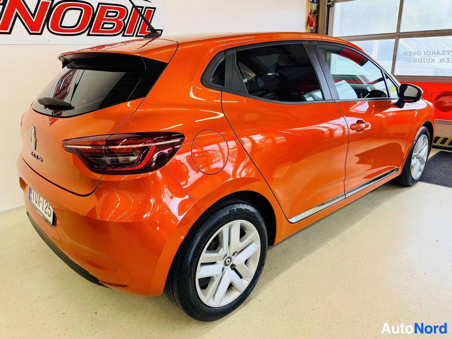 Takaviisto: Oranssi Renault Clio paikallaan varikolla, kuvattu takaviistosta oikealta näkyen. Näkyvät takalamppu, pyörä ja sivupaneeli.