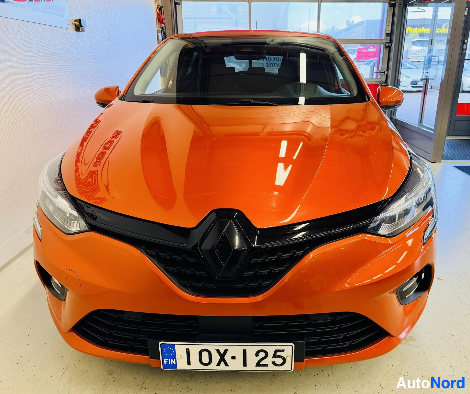 Edestä: Oranssi Renault Clio edestä kuvattu varikolla. Näkyvät sälöikkö, ajovalot, Renault-logo ja rekisterikilpi ''