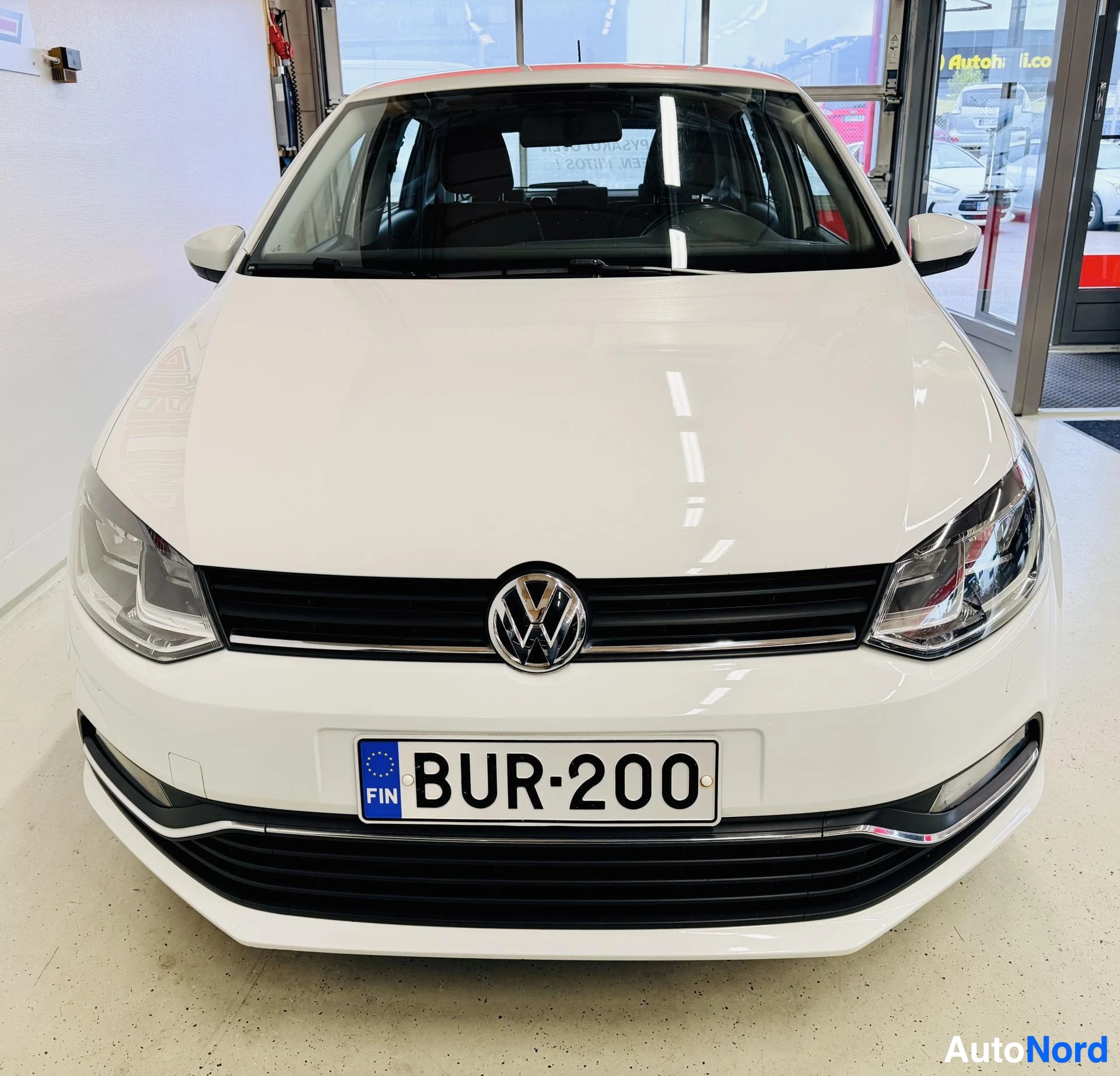 Edestä: Valkoinen Volkswagen Polo edestä katsottuna, VW-logo keskellä, puolipyöreät ajovalot, rekisterikilpi, sisätiloissa