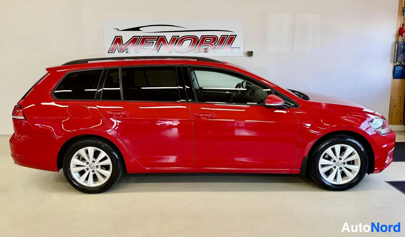 Sivulta: Punainen Volkswagen Golf Variant, sivunäkymä vasemmalle, sisätiloissa pysäköity. Ajoneuvon koko pituus näkyvissä, valkopalliset vanteet ja harmaat ikkunasävyt.