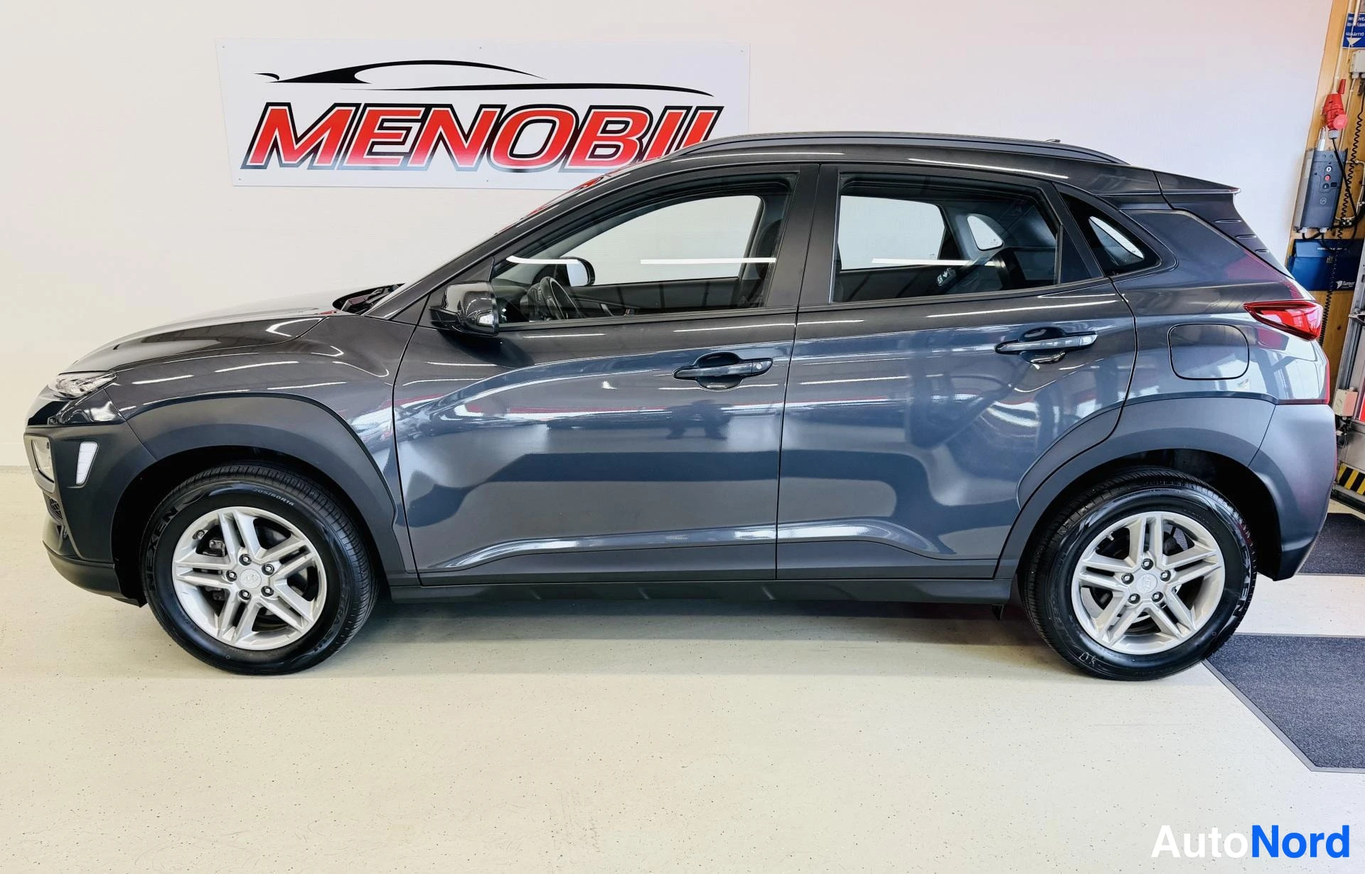 Sivulta: Tummansininen Hyundai Kona puhtaasta sivusta, sisätiloissa olevassa showroom-hallissa, valkoisilla vanteillla, ovet auki.