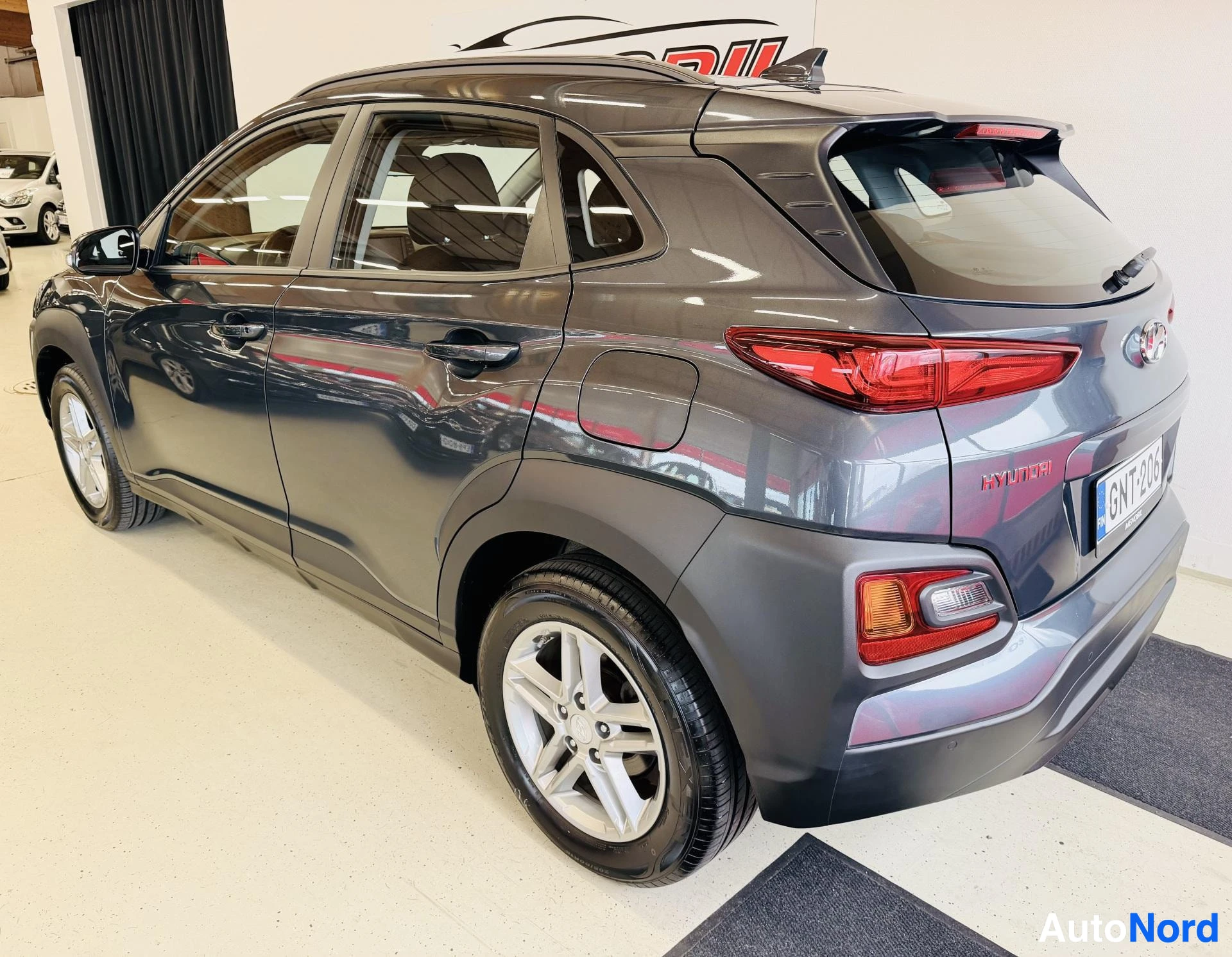 Takaviisto: Tummansininen Hyundai Kona takapuolen kolmen neljänneksen näkymässä, ovet auki, rekisterinumero GNT206 näkyy, valkoiset vanteet, punainen silmäys.
