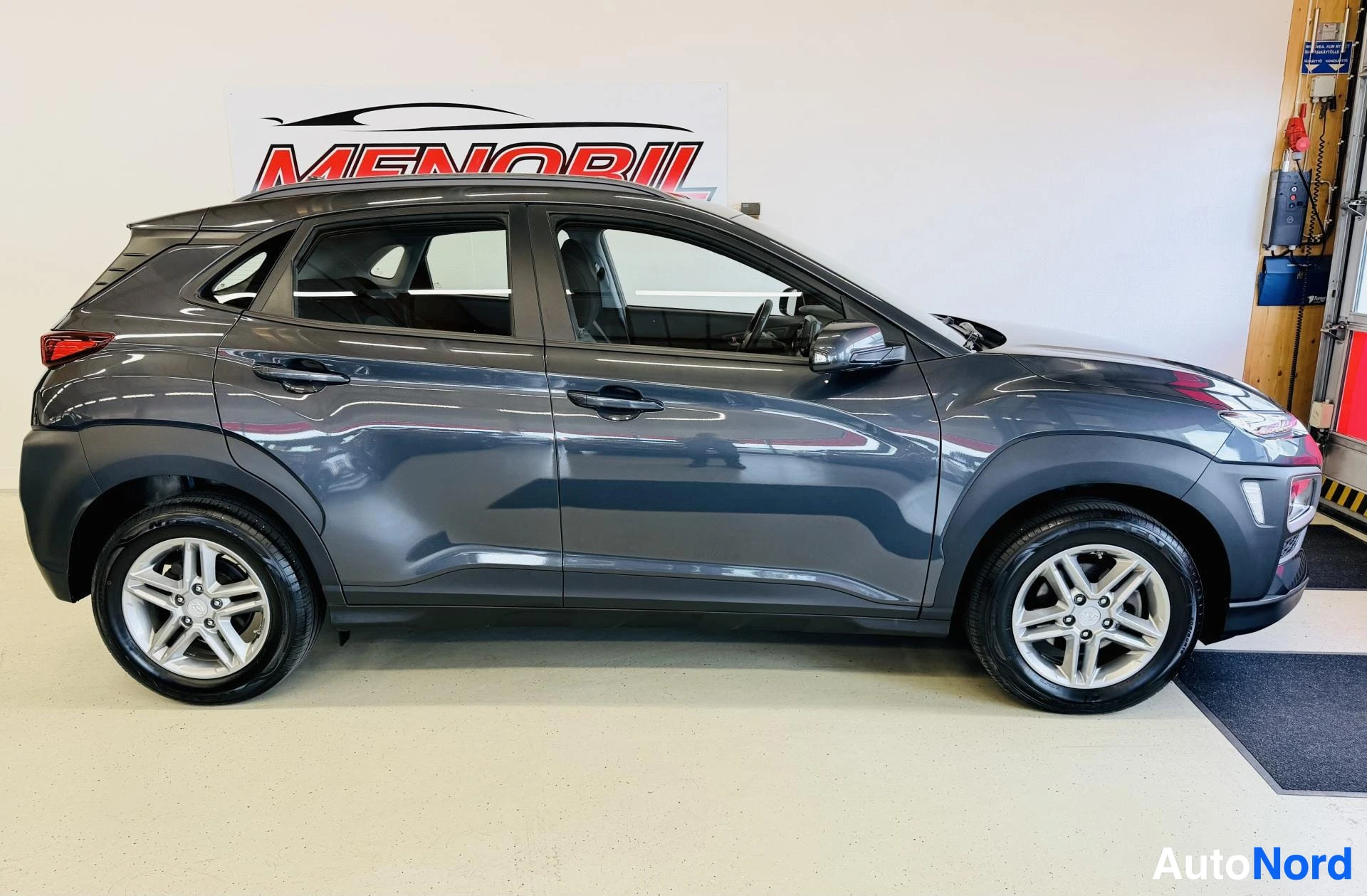 Sivulta: Harmaa Hyundai Kona näyttelyhallissa, kuvattu sivulta vasemmalta. Ajoneuvolla valkoiset alumiinivanteet ja mustat ikkunat.