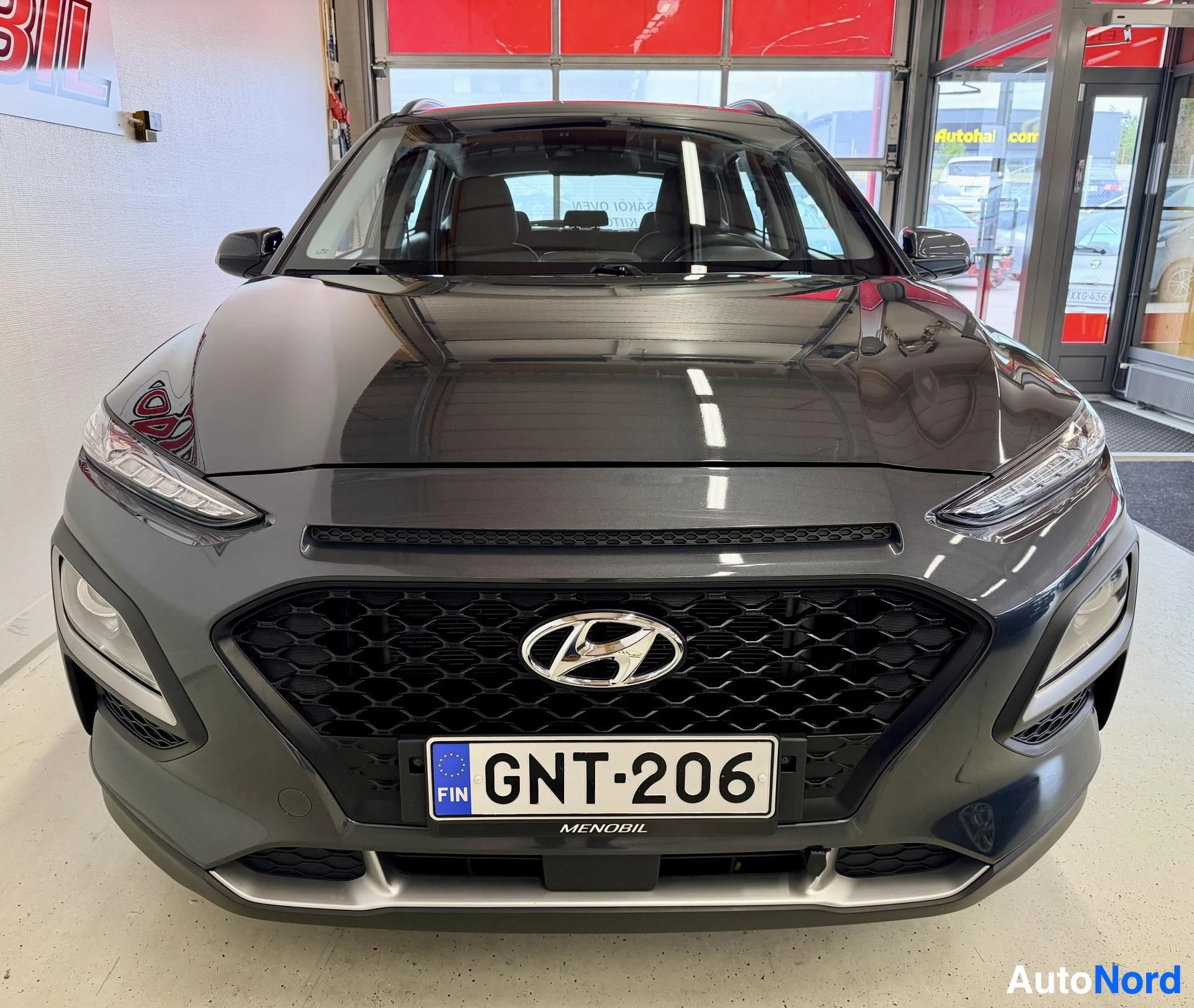 Edestä: Harmaa Hyundai Kona kuvattu edestä näyttelyhallissa. Rekisterikilvessä, Hyundai-logo keskellä grillissä
