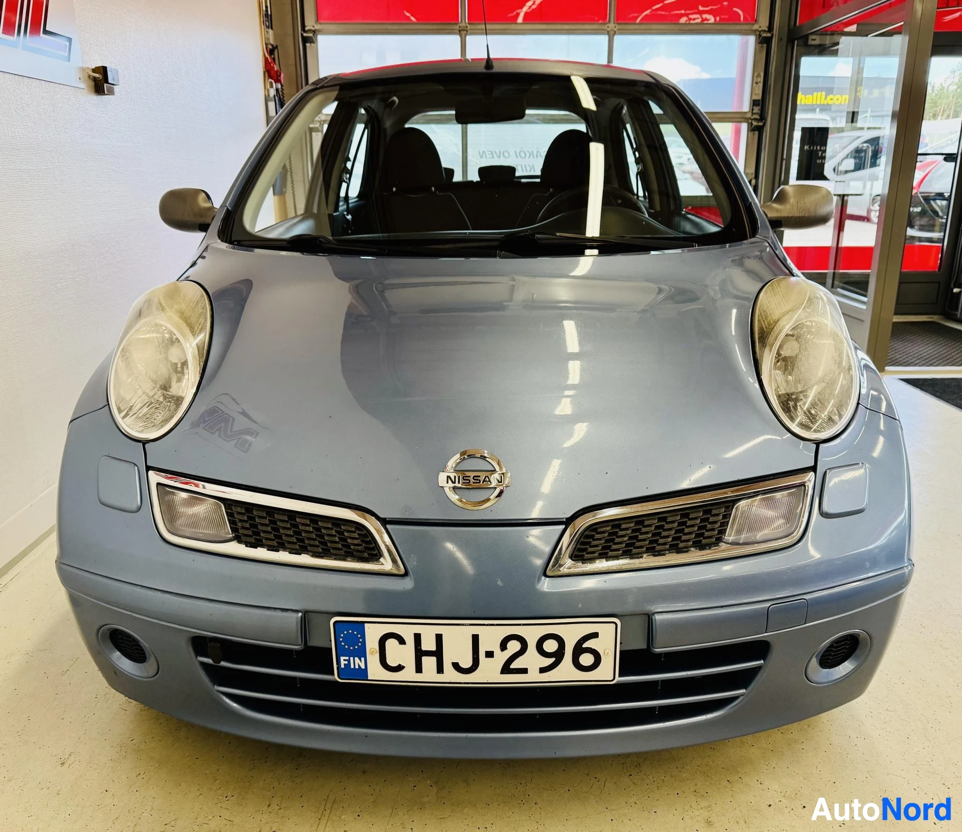 Edestä: Nissan Micra etuosa, hopea väri, kahdet kuplavalot, Nissan-tunnus keskellä, tumma grilli, rekisterikilvessä CHJ296, tumma lasitus, sisätila näkyvissä.